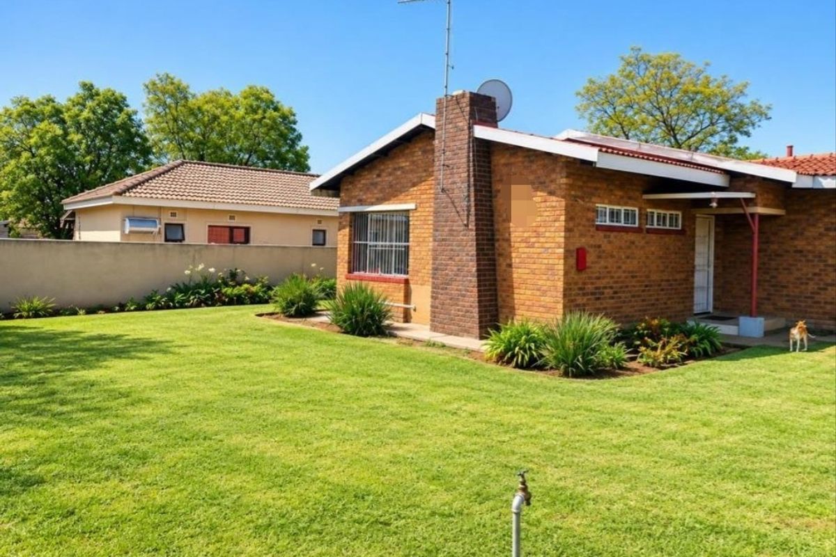 A view of the rear exterior of a brick house featuring a chimney, tiled roof, and a well-maintained lawn with a boundary wall.