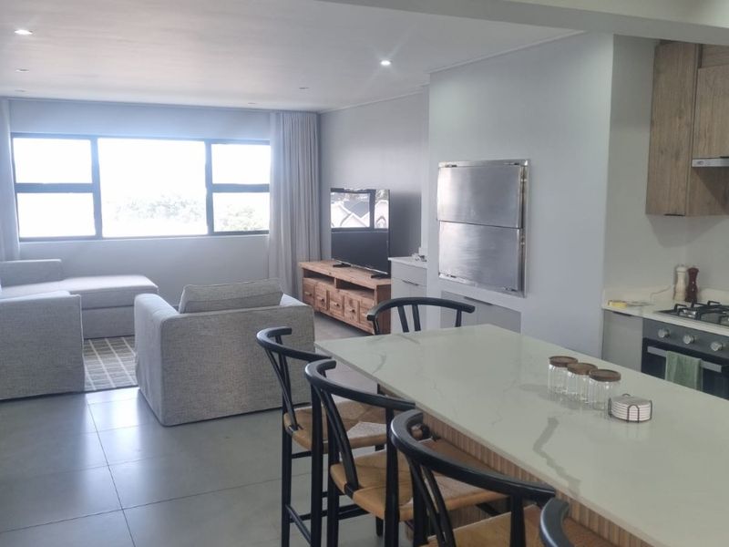 Modern open-plan living area featuring a kitchen island with stone countertops, built-in braai, and tiled flooring, seamlessly connecting to the lounge space.