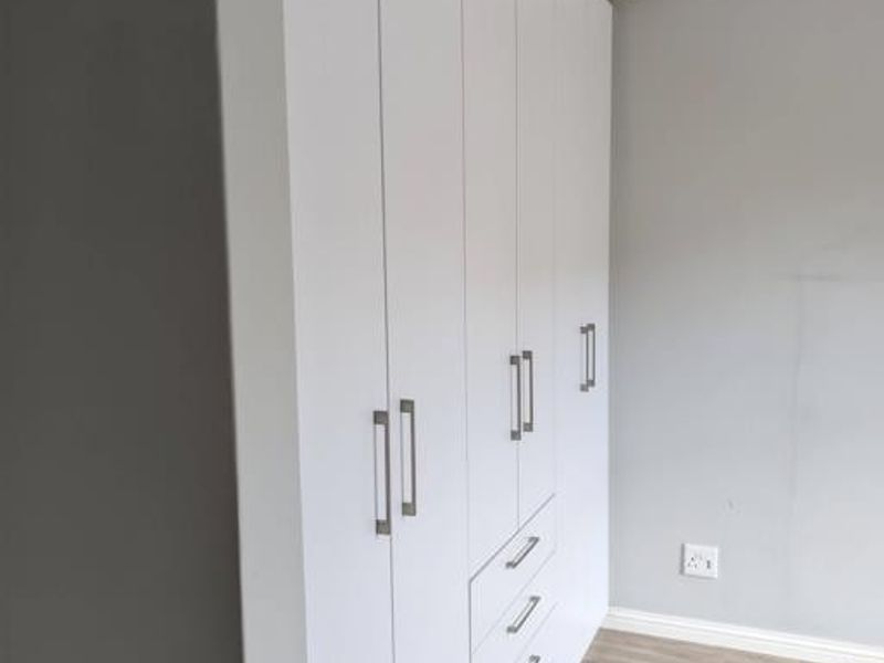 A clean, modern bedroom featuring extensive white built-in cupboards, light-toned laminate flooring, and recessed ceiling lighting.