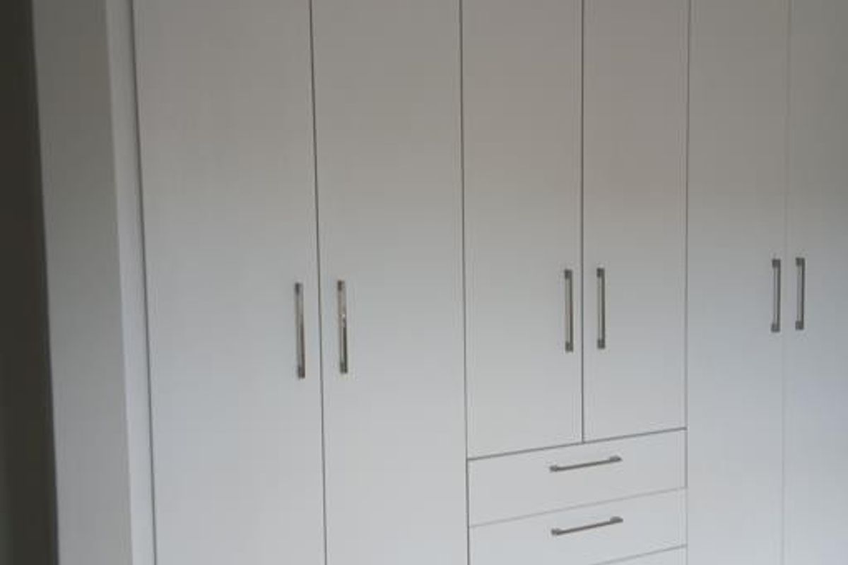 A clean, modern bedroom featuring floor-to-ceiling white built-in cupboards with integrated drawers and light-toned laminate flooring.