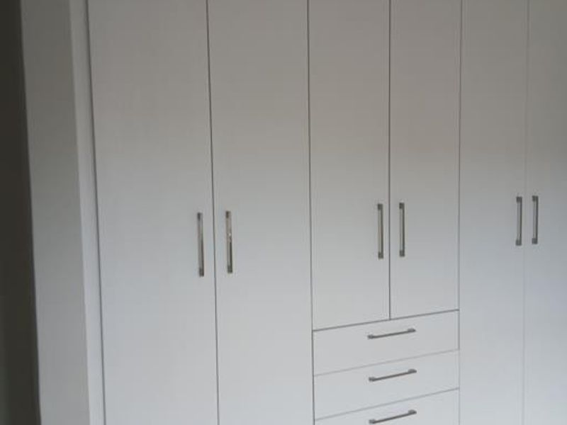 A clean, modern bedroom featuring floor-to-ceiling white built-in cupboards with integrated drawers and light-toned laminate flooring.