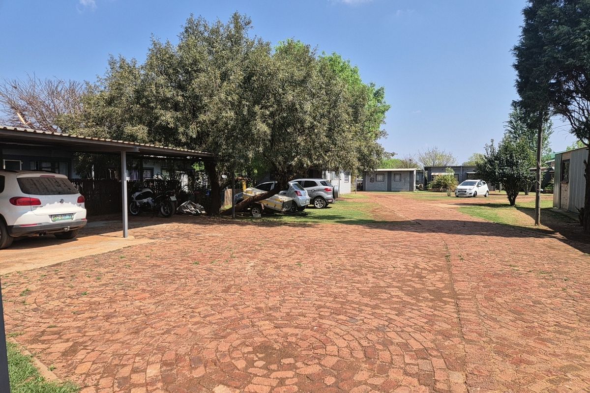 Spacious industrial property exterior featuring a large paved driveway, covered carport area, and established garden surroundings.
