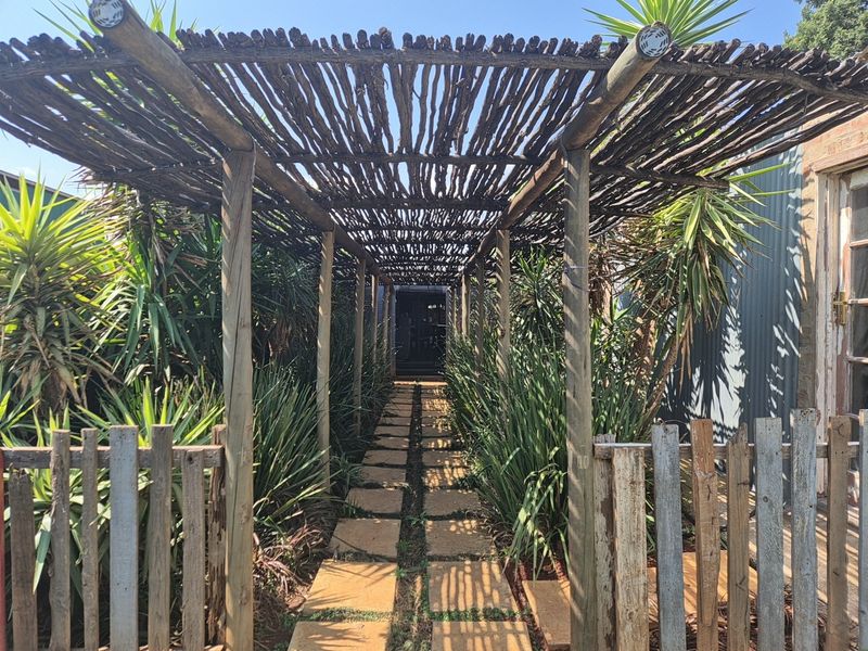A rustic entrance featuring a wooden pergola walkway with stone pavers, framed by a picket fence and established garden landscaping.