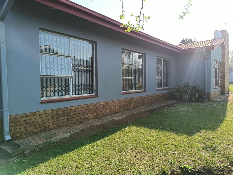 Rear exterior view of the house featuring a grey painted facade, brick plinth, and secure metal window gates overlooking a lawn area.