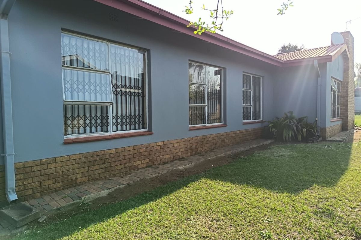 Rear exterior view of the house featuring a grey painted facade, brick plinth, and secure metal window gates overlooking a lawn area.