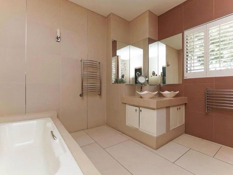 A spacious bathroom featuring a built-in bathtub, double vanity with integrated sinks, heated towel rails, and plantation shutters for natural light control.