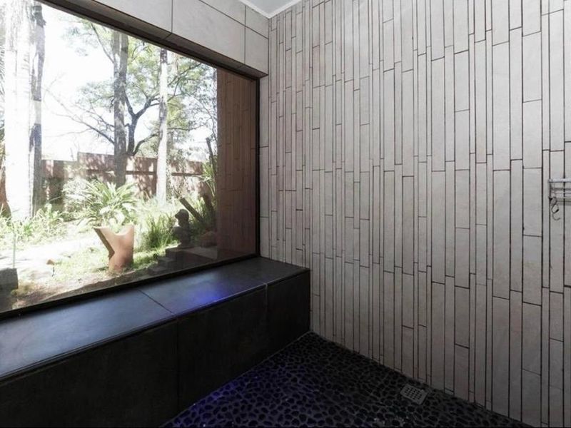 A modern bathroom featuring a large window with garden views, textured wall tiling, and a built-in bench with a pebble-style floor.