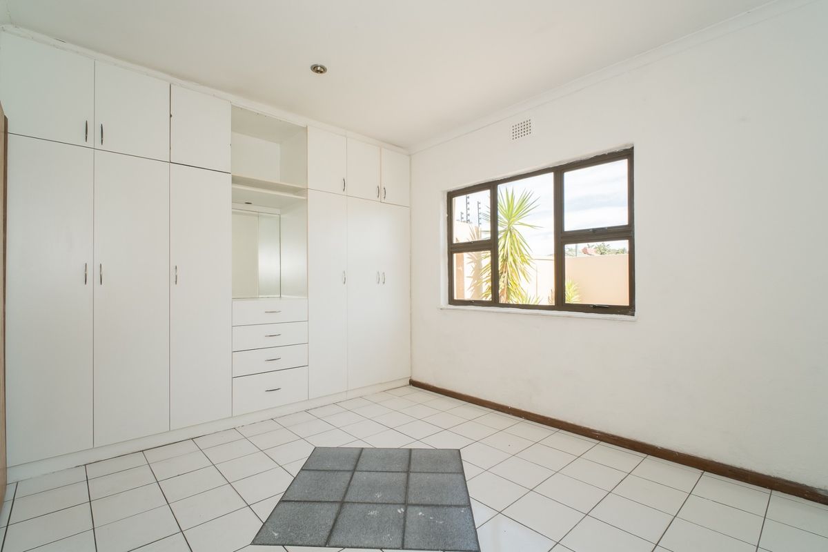 A bright, empty bedroom featuring white built-in cupboards, tiled flooring, and a large window providing natural light.