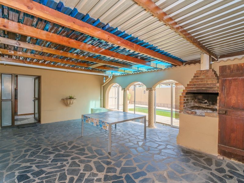 Covered outdoor patio featuring a built-in brick braai, stone flooring, and timber beam roof structure, overlooking the pool area.