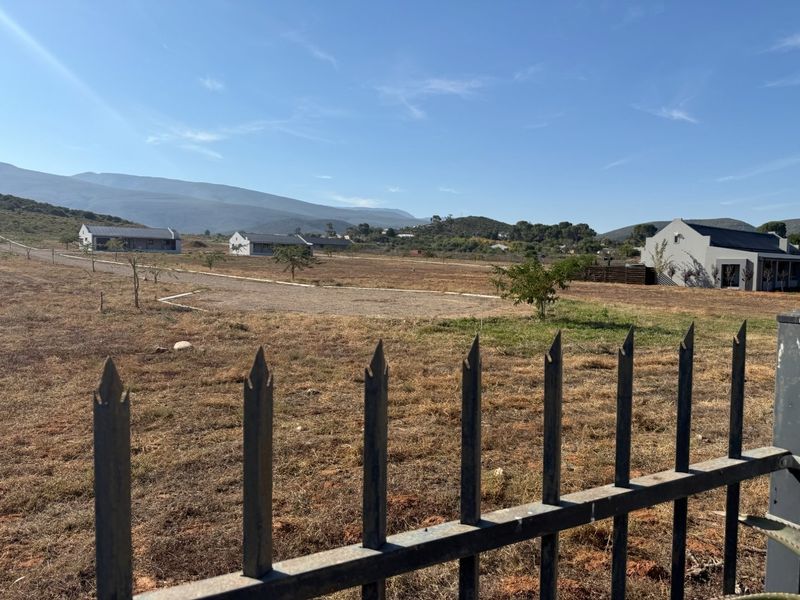 A view of a vacant residential plot featuring metal perimeter fencing, an unpaved driveway, and surrounding residential buildings with mountain views.