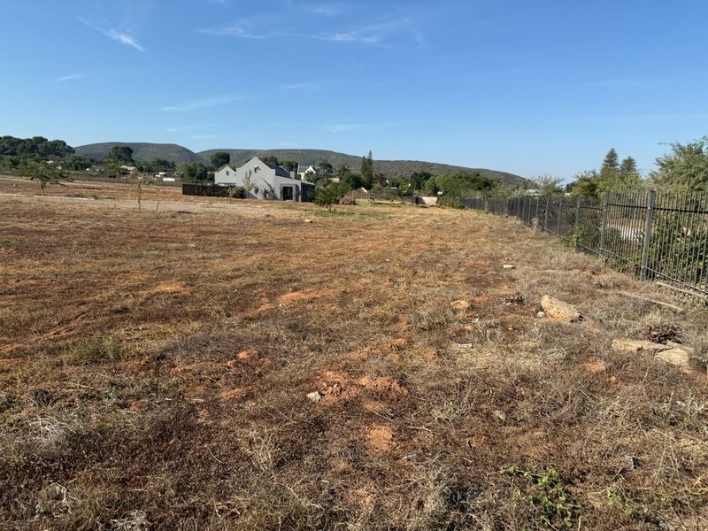 A view of a vacant residential plot featuring dry grass and metal perimeter fencing, with surrounding landscape and distant buildings visible.