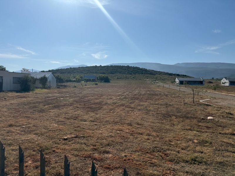 A wide-angle view of a vacant residential plot featuring dry grass, perimeter fencing, and surrounding mountain views in a quiet rural setting.