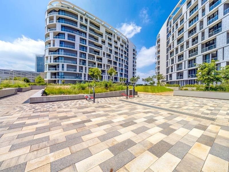 A well-maintained paved courtyard area between modern apartment buildings, featuring landscaped garden beds and outdoor fitness equipment.
