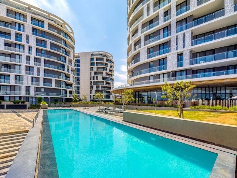 A modern apartment complex featuring a pristine swimming pool, landscaped garden areas, and contemporary architectural design with glass-fronted balconies.