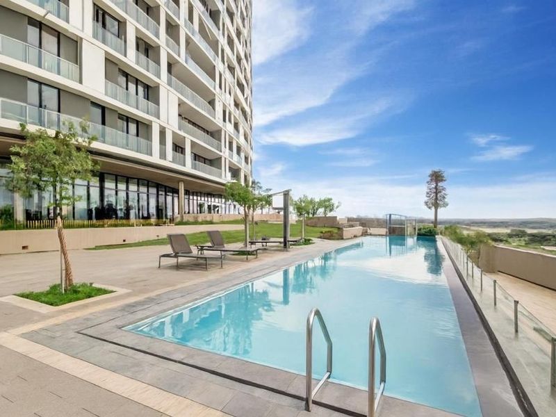 A modern, well-maintained swimming pool area featuring a paved deck, glass balustrades, and scenic views, situated adjacent to the apartment building.