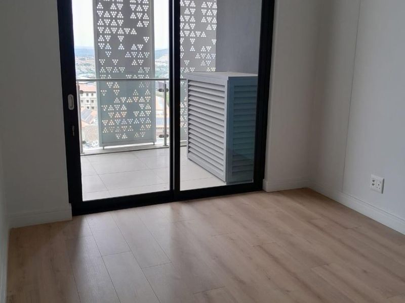 A bright, empty bedroom featuring modern laminate flooring and a sliding glass door leading to a private balcony with decorative privacy screens.