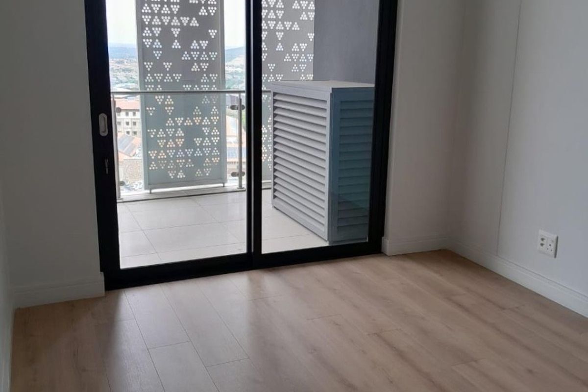A bright, empty bedroom featuring modern laminate flooring and a sliding glass door leading to a private balcony with decorative privacy screens.