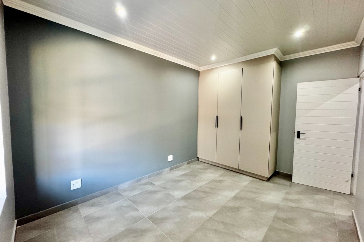 A clean, modern bedroom featuring neutral tiled flooring, built-in cupboards, and a stylish tongue and groove ceiling with recessed lighting.