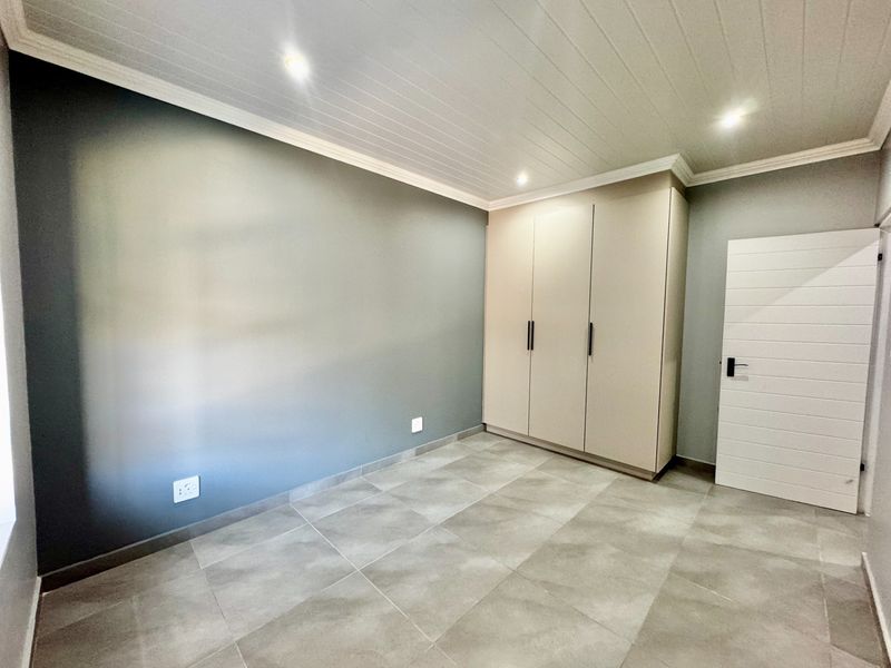 A clean, modern bedroom featuring neutral tiled flooring, built-in cupboards, and a stylish tongue and groove ceiling with recessed lighting.