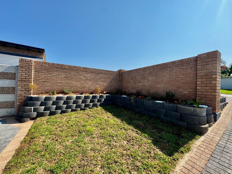 A well-maintained garden area featuring a brick boundary wall, tiered retaining walls, and a neat lawn adjacent to a paved driveway.