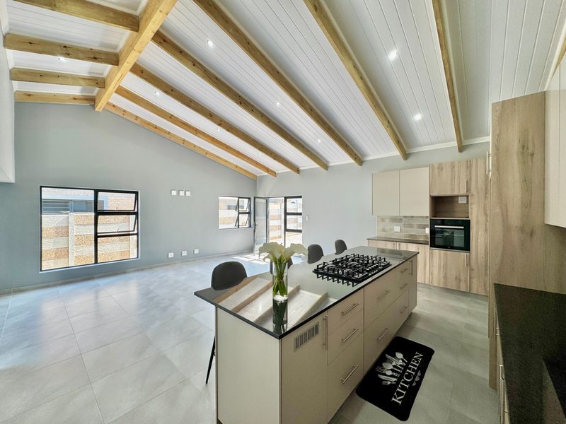 Modern open-plan living area featuring a vaulted ceiling with exposed timber beams, tiled floors, and a contemporary kitchen with a central island and gas hob.