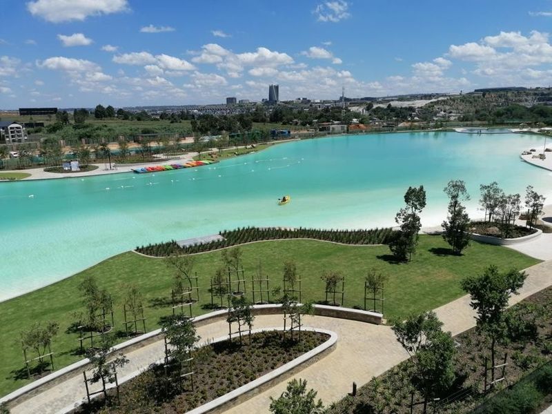 Elevated view overlooking a large turquoise man-made lagoon, landscaped green spaces, and paved pathways within a modern residential estate.