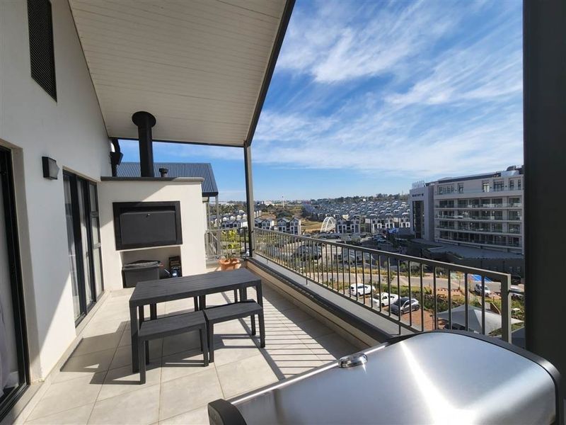 Spacious covered balcony featuring a built-in braai, tiled flooring, and metal railings, offering expansive views of the surrounding estate.