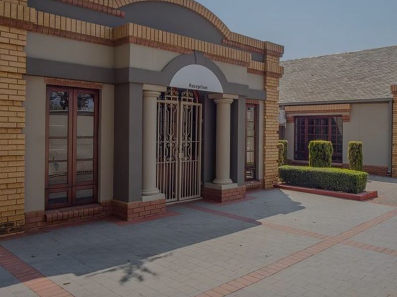 Front entrance facade featuring brick detailing, decorative columns, and a secure metal gate, leading to a paved driveway area.
