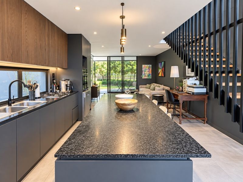 Modern open-plan kitchen and living area featuring a large stone island, sleek dark cabinetry, integrated appliances, and a striking staircase with vertical balustrades.