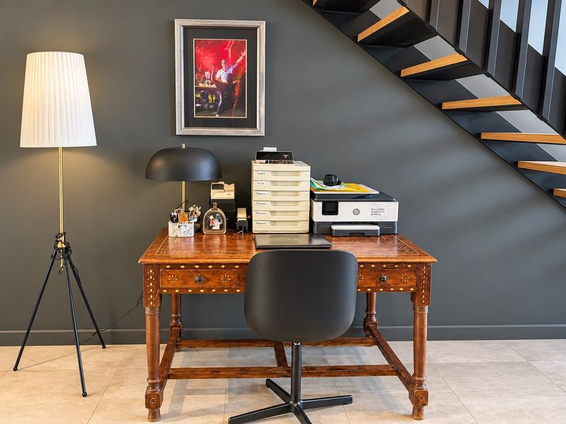 A dedicated home office space featuring modern open riser staircase design and neutral tiled flooring against a dark feature wall.