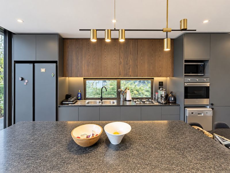 Modern kitchen featuring dark grey cabinetry, wood-grain upper cabinets, stone countertops, integrated appliances, and a central island with contemporary pendant lighting.