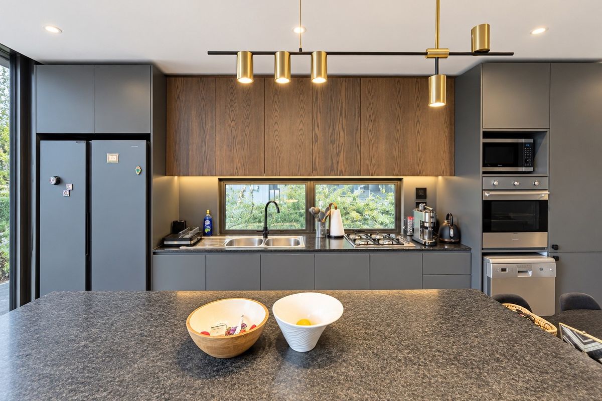 Modern kitchen featuring dark grey cabinetry, wood-grain upper cabinets, stone countertops, integrated appliances, and a central island with contemporary pendant lighting.