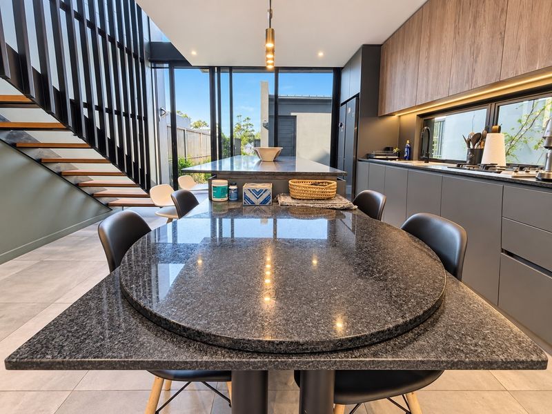 Modern open-plan kitchen and dining area featuring a large stone island, sleek cabinetry, and a striking staircase, opening onto an outdoor space.