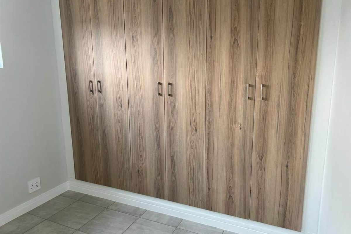 A clean, modern bedroom featuring floor-to-ceiling wood-grain built-in cupboards and neutral tiled flooring.