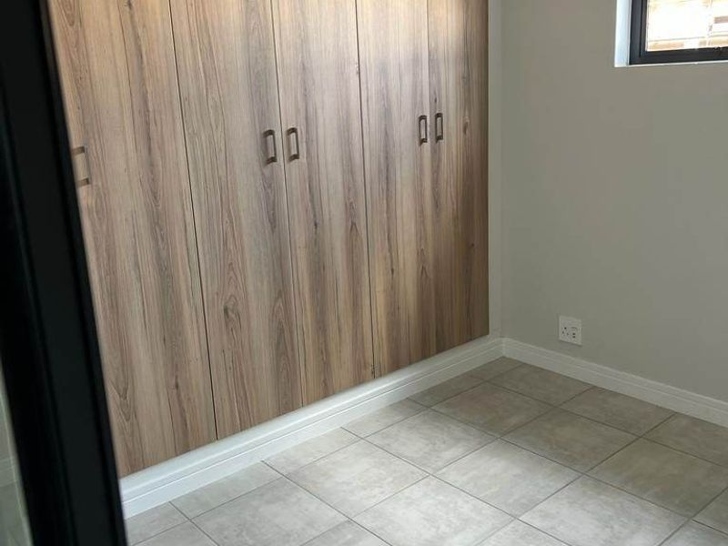 A clean, modern bedroom featuring neutral tiled flooring, light grey walls, and extensive floor-to-ceiling wood-finish built-in cupboards.