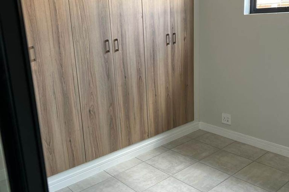 A clean, modern bedroom featuring neutral tiled flooring, light grey walls, and extensive floor-to-ceiling wood-finish built-in cupboards.