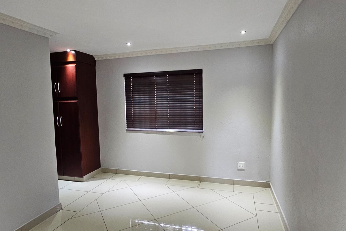 A clean, empty bedroom featuring neutral tiled flooring, recessed downlighting, decorative ceiling cornicing, and a built-in dark wood cupboard.