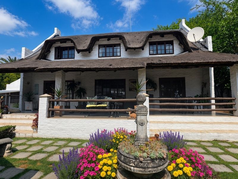 A charming rear elevation of a thatched-roof house featuring a spacious covered patio, a stone fountain, and a well-maintained garden path.
