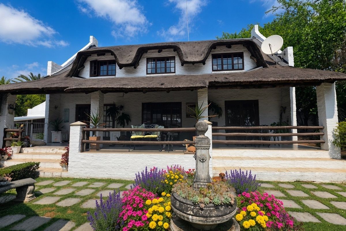 A charming rear elevation of a thatched-roof house featuring a spacious covered patio, a stone fountain, and a well-maintained garden path.