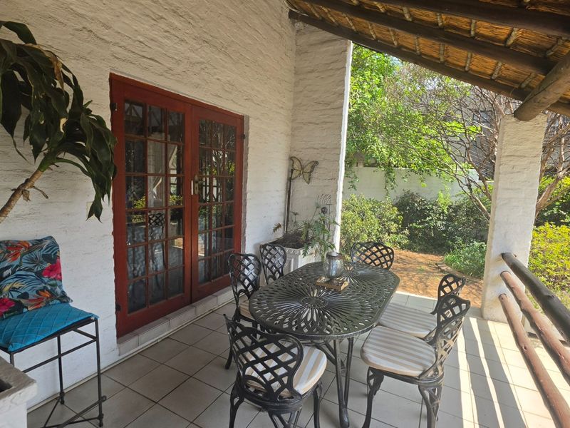 A covered patio area featuring a thatched roof, tiled flooring, and timber-framed glass doors leading into the home, overlooking a garden space.