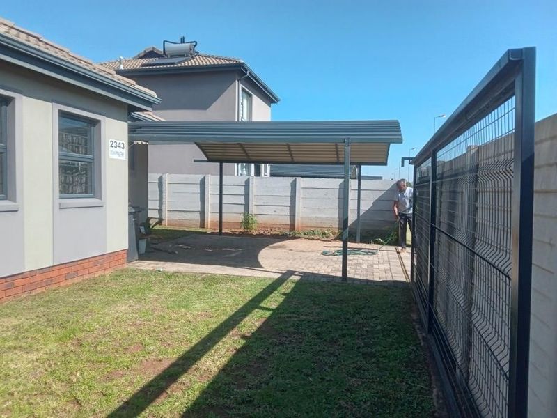 Front exterior view featuring a paved driveway, covered carport, mesh perimeter fencing, and a sliding gate, with a solar water heater installed on the roof.