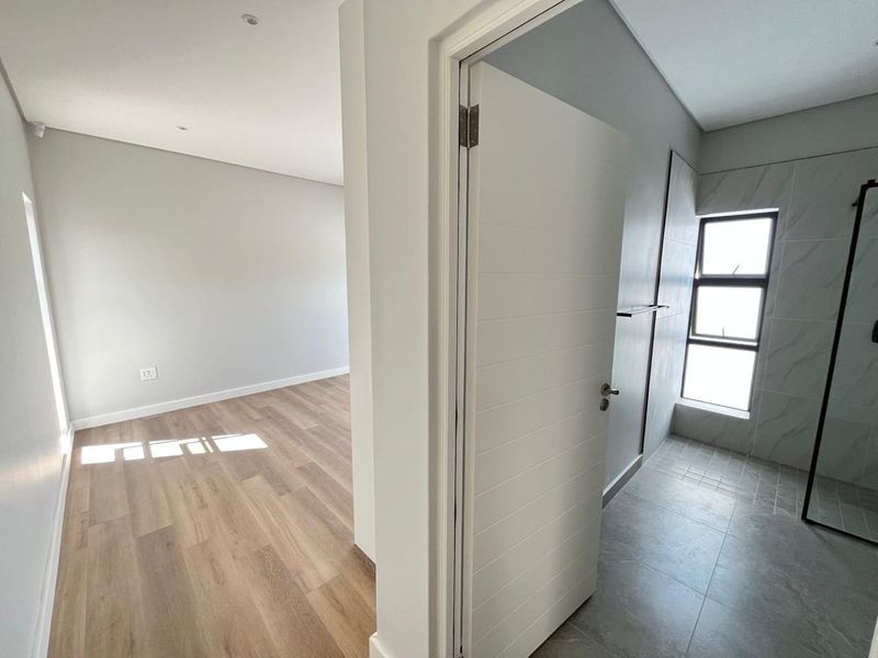 A bright, modern hallway transition area connecting a bedroom with wood-look flooring to a contemporary bathroom featuring tiled finishes and a glass shower enclosure.