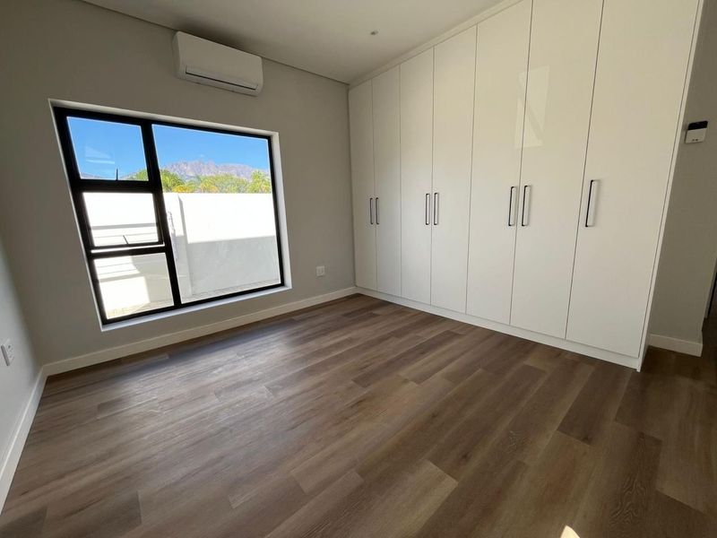 A bright, modern bedroom featuring sleek built-in cupboards, durable laminate flooring, and a wall-mounted air conditioning unit with views of the surrounding landscape.