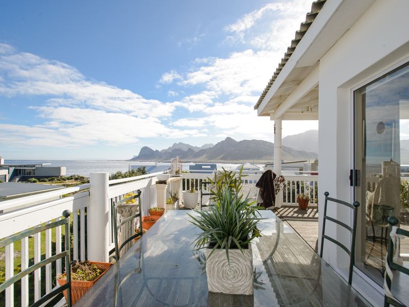 Spacious elevated balcony featuring wooden decking, white balustrades, and expansive coastal and mountain views, accessible via sliding glass doors.