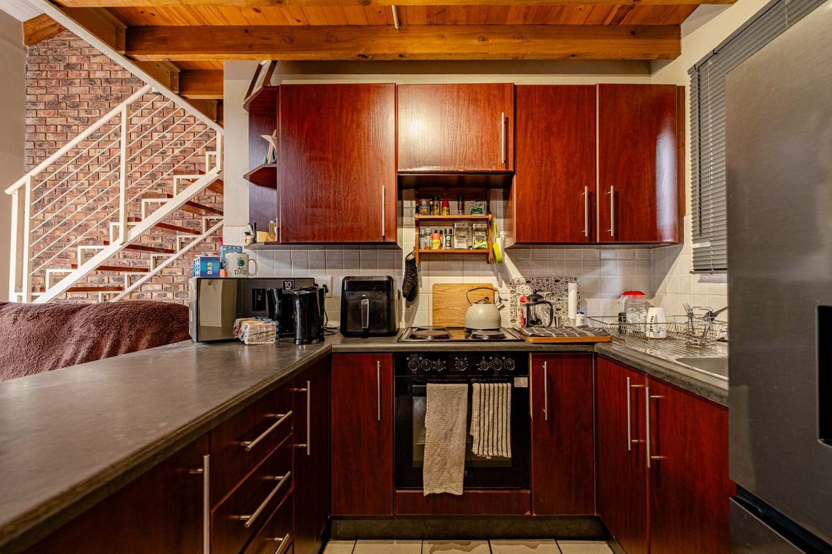 Compact kitchen featuring dark wood cabinetry, laminate countertops, and an integrated oven and hob, set against an exposed brick wall and timber ceiling beams.