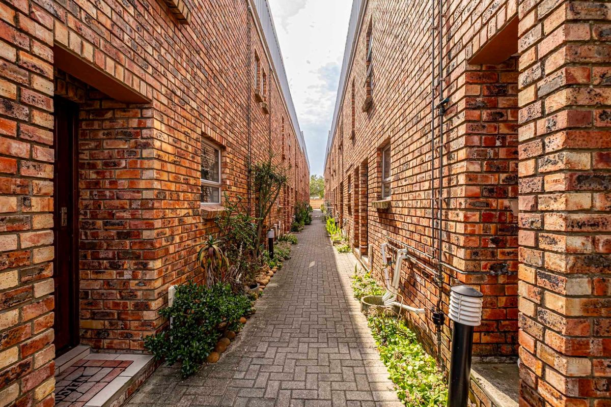 A narrow paved walkway between two exposed brick apartment blocks, featuring external lighting and established garden beds along the building perimeter.