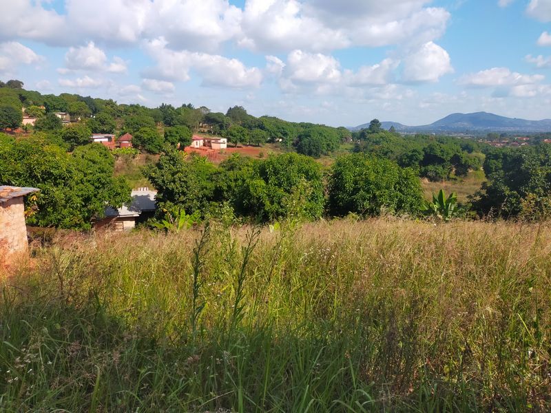 A scenic view overlooking a sloped, grassy residential plot with surrounding lush greenery and distant mountain vistas.
