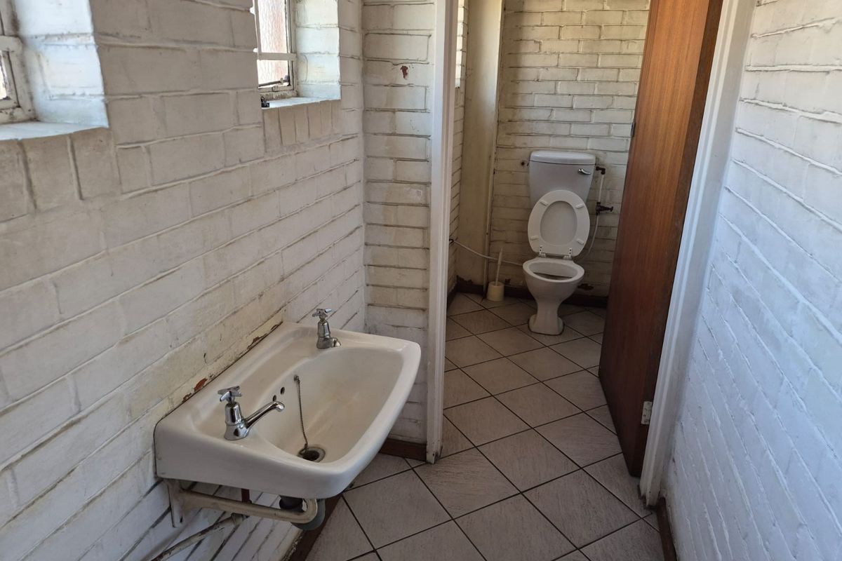 A functional bathroom featuring a wall-mounted basin, ceramic toilet, tiled flooring, and painted brick walls with security bars on the windows.
