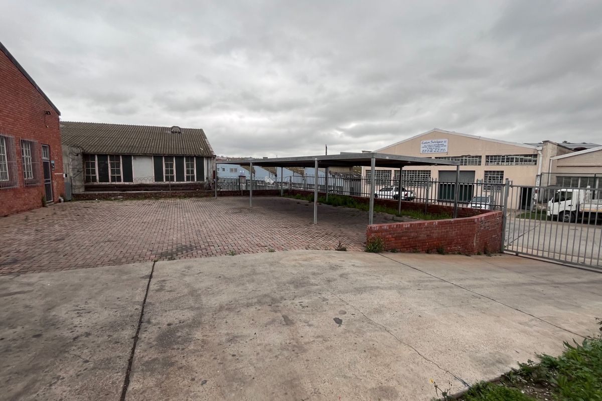 Industrial rear yard featuring brick paving, a covered carport structure, and secure metal perimeter fencing with concrete driveway access.