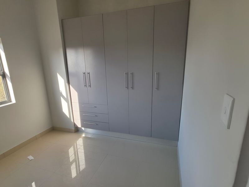 A bright, empty bedroom featuring modern built-in cupboards, neutral tiled flooring, and a large window providing natural light.